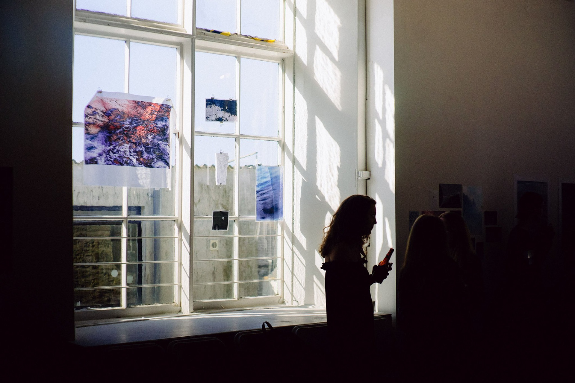 Gallery image of a window displaying Katie Riesner's work "A Journey Within". These works are printed on fabric and hung using fishing wire and bulldog clips, looking suspended midair. The printed photographs represent Riesner's feelings expressed outwardly through photo-manipulation of a red and blue rough sea, a bird that could either be seen as flying off, or landing and abstract imagery like a blue downwards spiral.