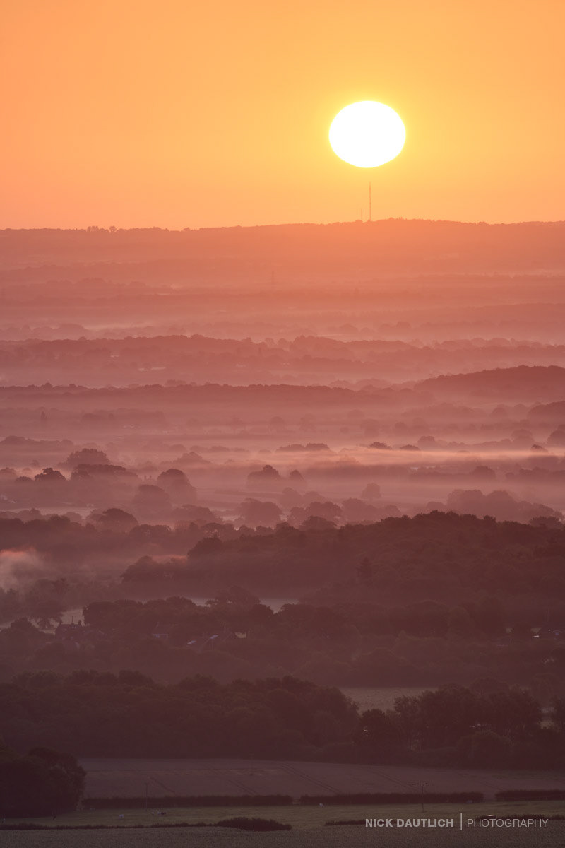 Landscape photo of mist hangs over Sussex hills and valleys during summer sunrise with bright sun in the sky
