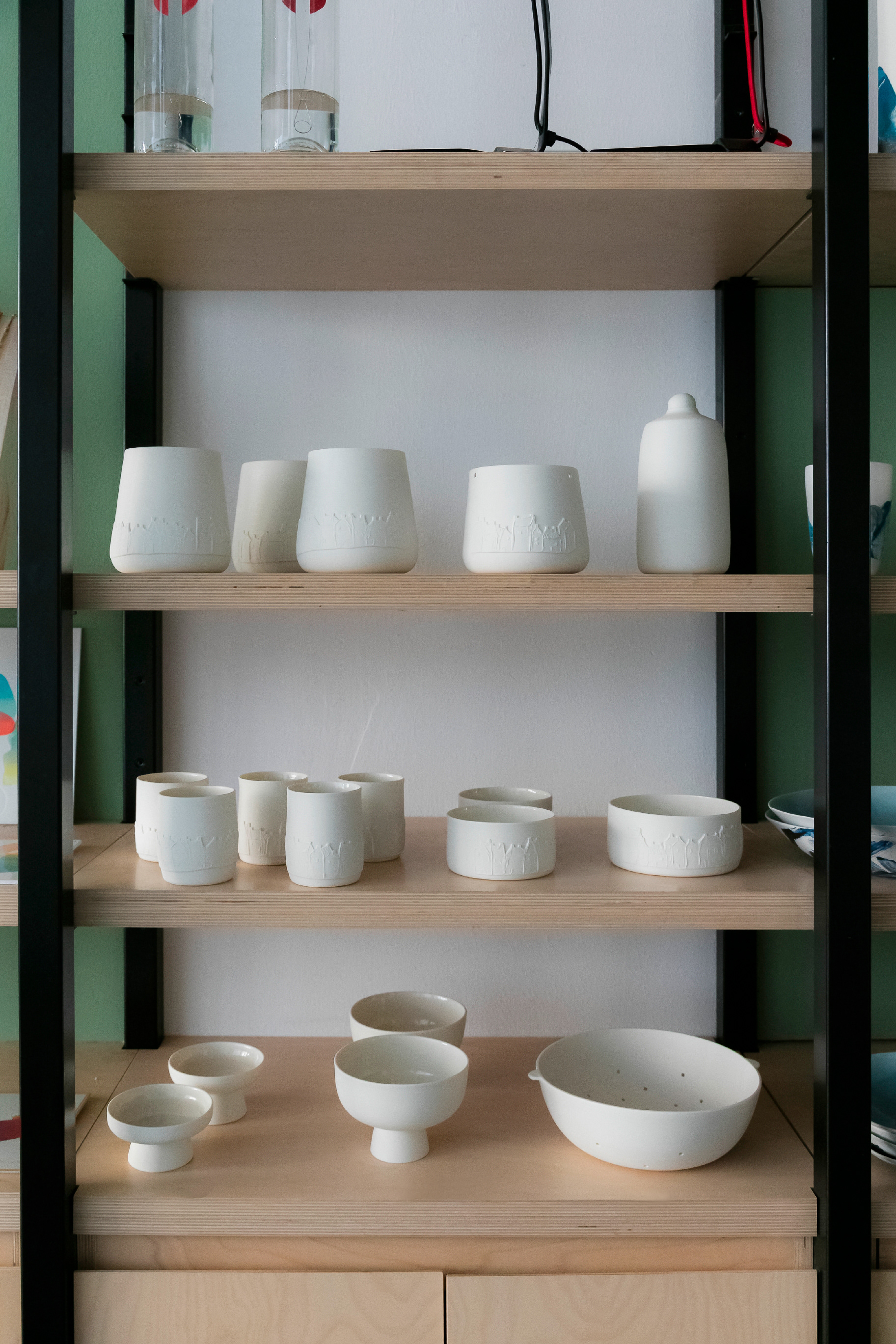 Collection en porcelaine blanche - De bas en haut: Passoire/Coupe à fruits 86€ / Coupe mini avec pied 25€ / Coupe bol avec pied 84€ / Plat petit et grand modèle 40€ & 50€ / Tasse avec decor maison 24€ / Bouteille avec bouchon 75€ / Pot petit et grand modèle avec decor 60€ & 65€