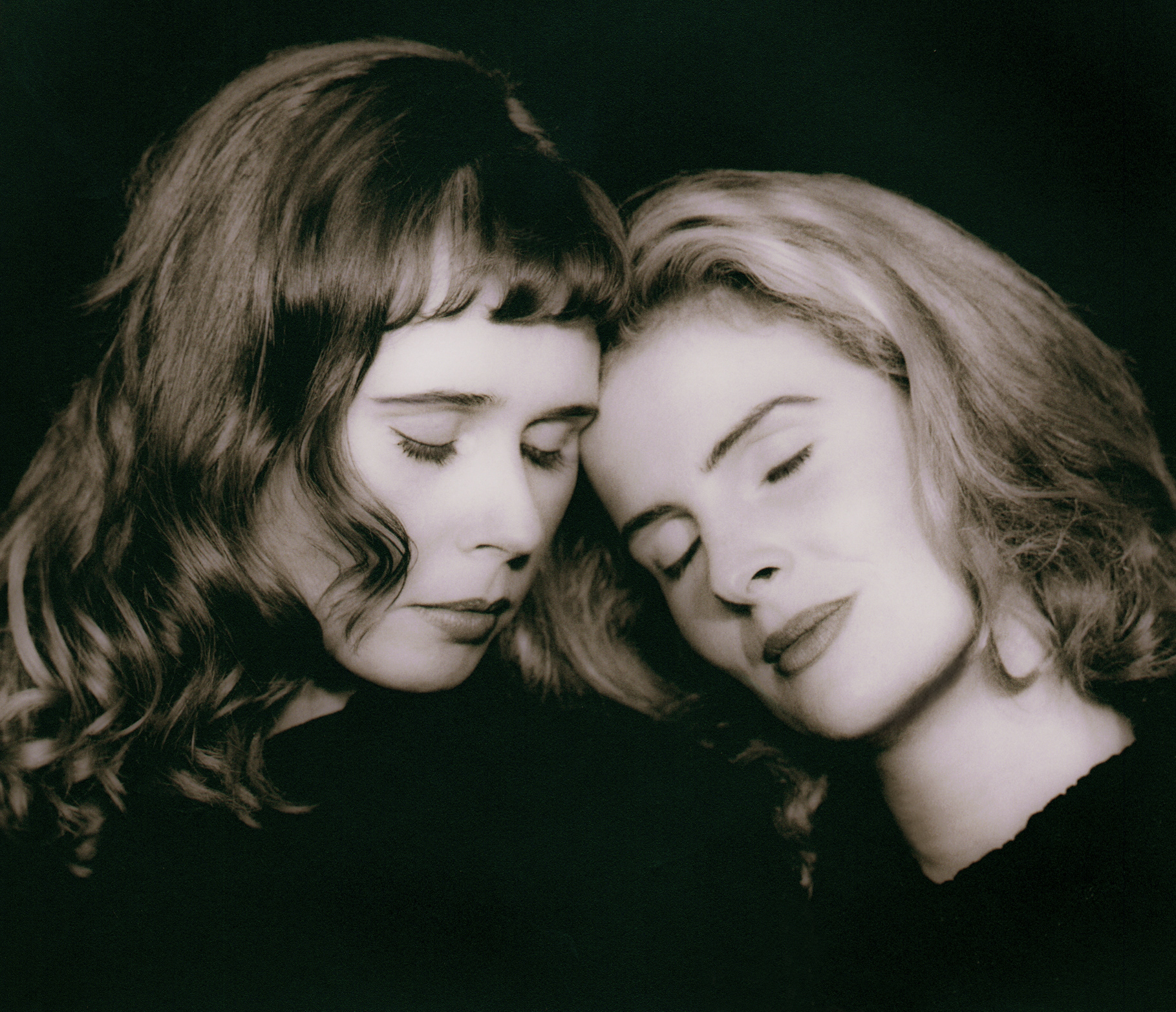 2 white womens faces leaning their heads together, eyes closed, against a black background in black and white