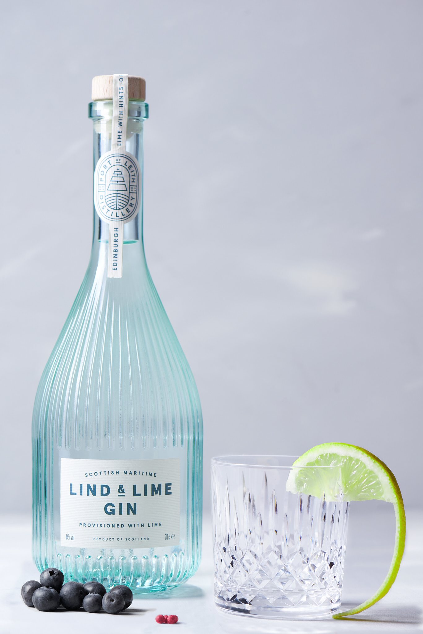 a bottle of Lind and Lime gin, brightly lit against a white, textured background. Next to it is a glass with a slice of lime placed on the rim. Around the bottle are small piles of pink peppercorns and juniper berries