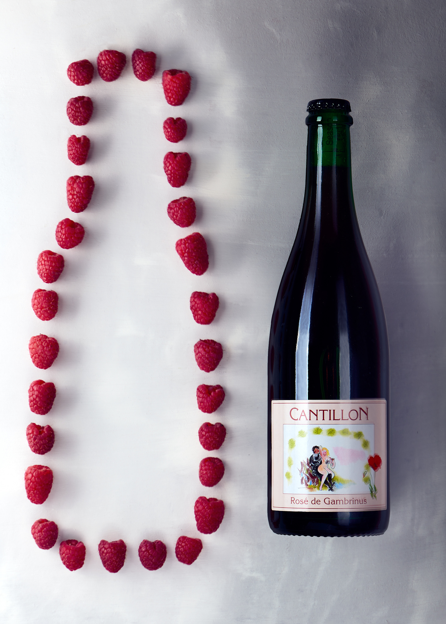 a flat-lay of a bottle of Cantillon Rose de Gambrinus on a light, texture background, with a light gradient from the left to the right. To the left of the bottle, raspberries are dotted in the outline of a bottle