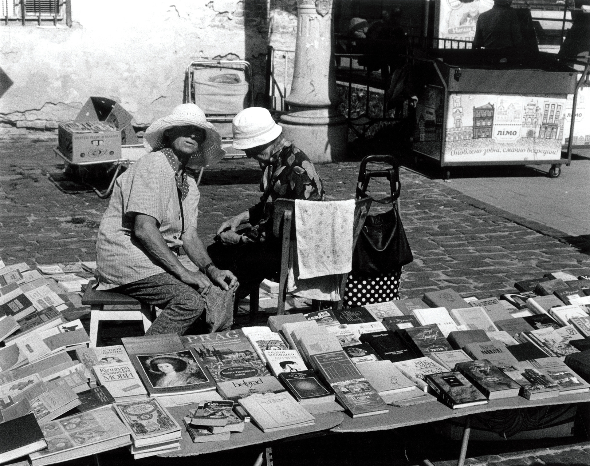2 women sit at their stall in the Lviv book market. One of the women looks quizzically at camera, the other pays no attention