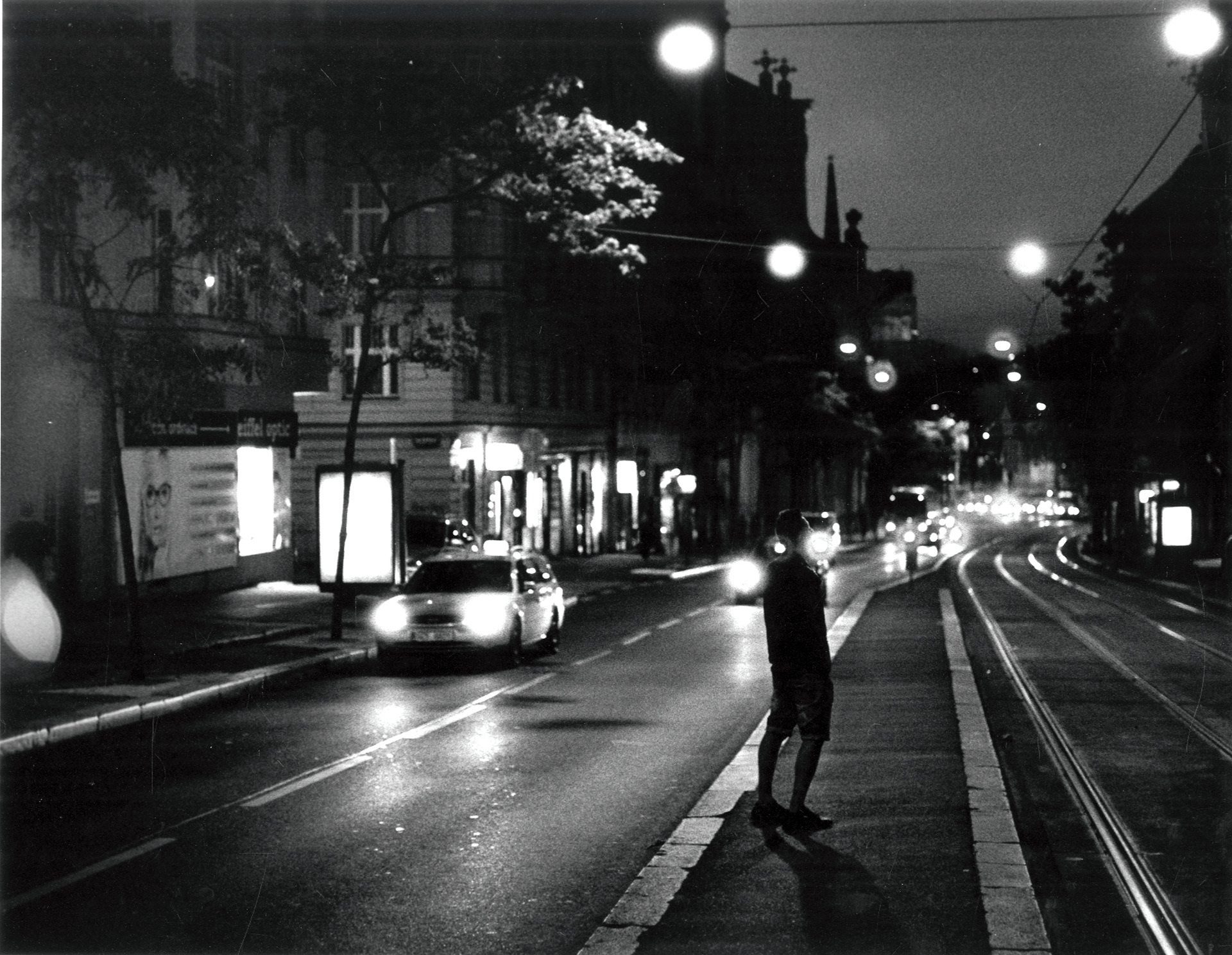 A man stands in the central reservation on a main road in Prague, backlit by passing cars' headlights