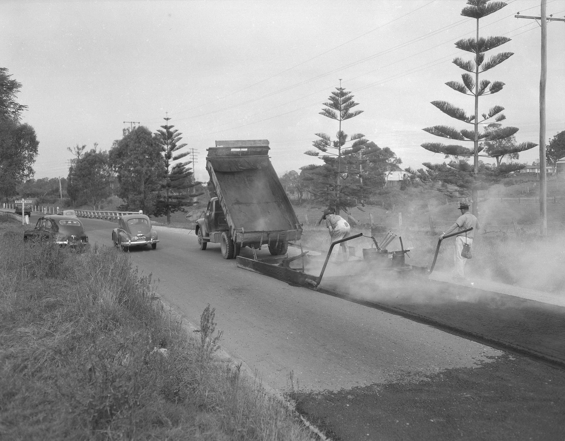 Redcliffe Road under construction. Two workers laying bitumen as it pours from the back of a truck