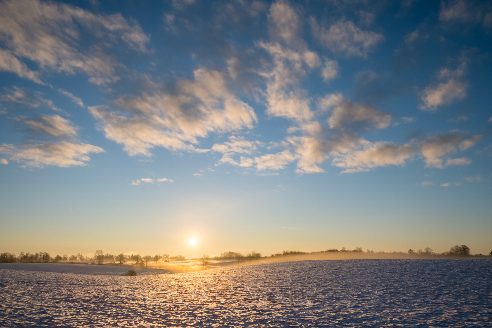 The rising Sun lights a dramatic sky of bright clouds above a cropped field under a light blanket of snow.
