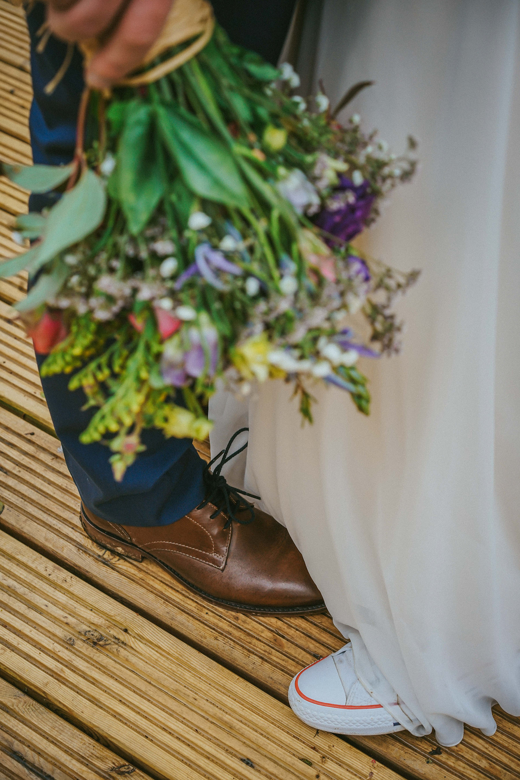 bouquet of colourful fresh wild flowers for wedding being held by groom with clear photo of brown smart shoes and white converse worn by bride