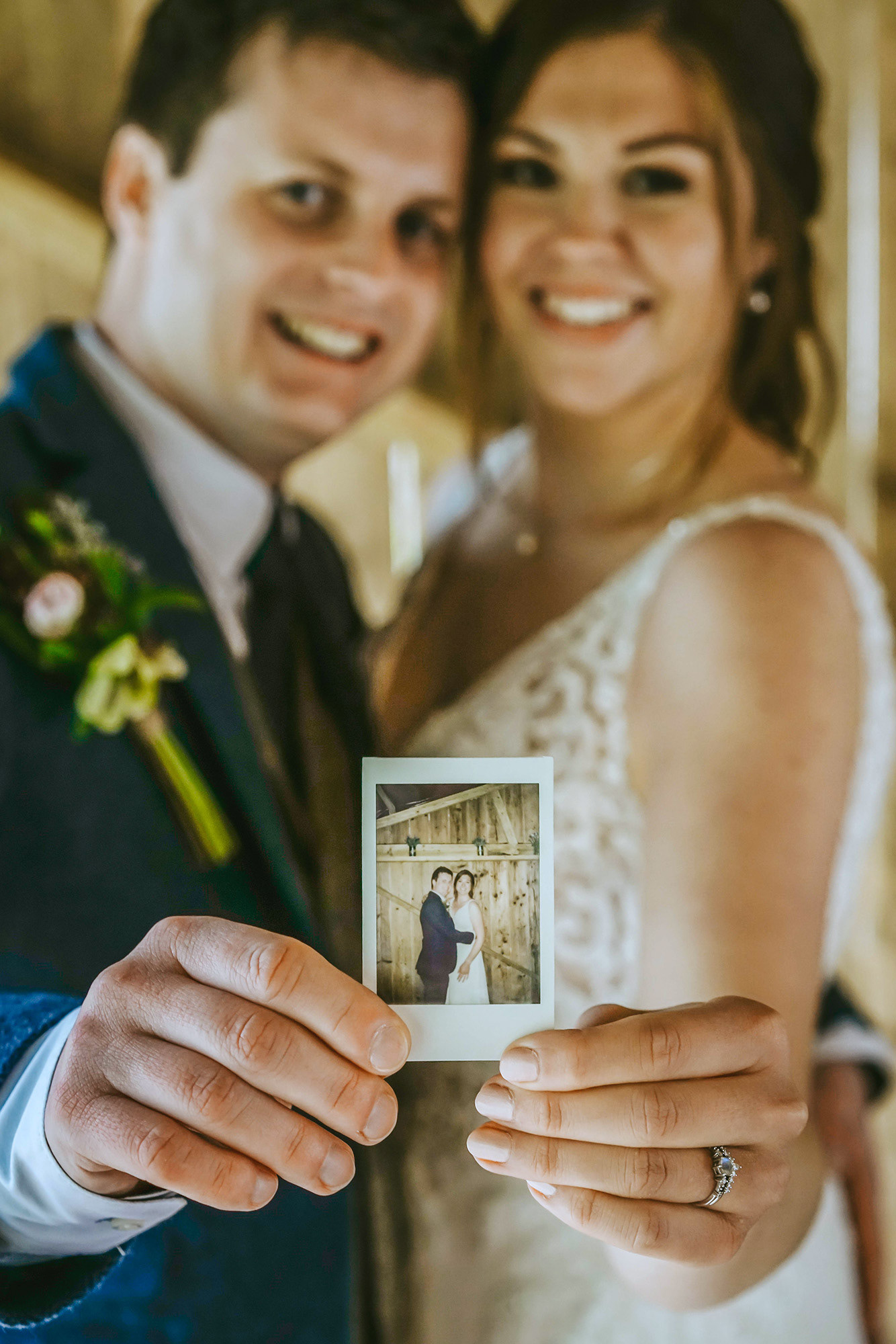 bride and groom smiling at photographer holding out an instant camera print out of them stood at wedding barn in buckinghamshire stood in classic wedding atire