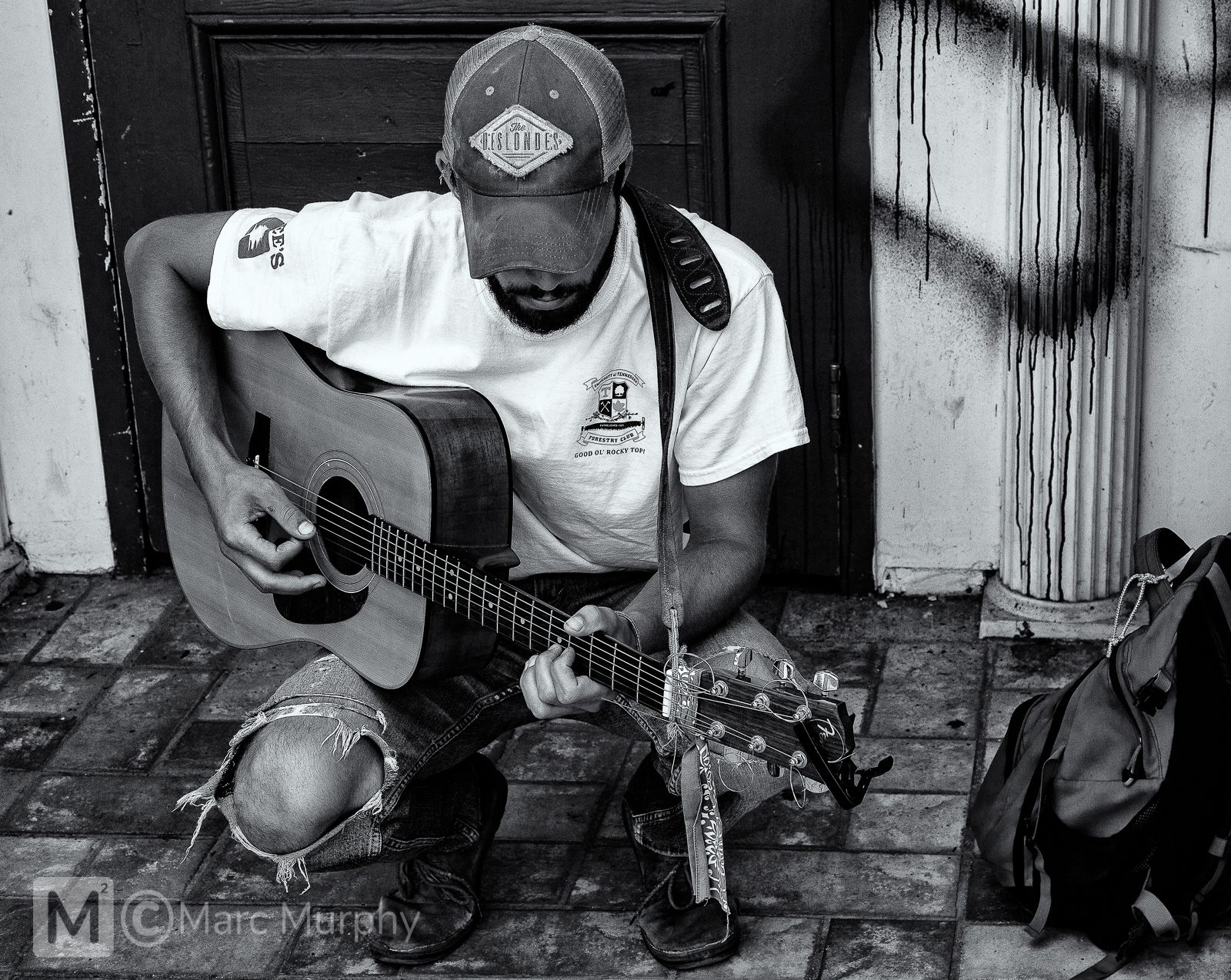 Black and White of Kneeling Street Musician Playing Guitar Wearing White T-Shirt, Old Trucker Hat and Ripped Blue Jeans