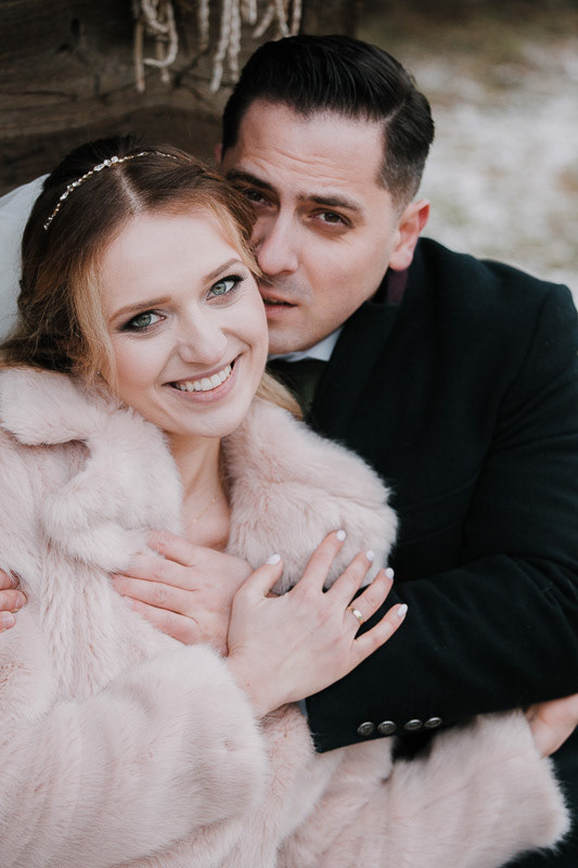 para młoda siedzi na ławce przytulając się, sesja zimowa pary młodej, panna młoda nosi futerko, fotografia ślubna białystok