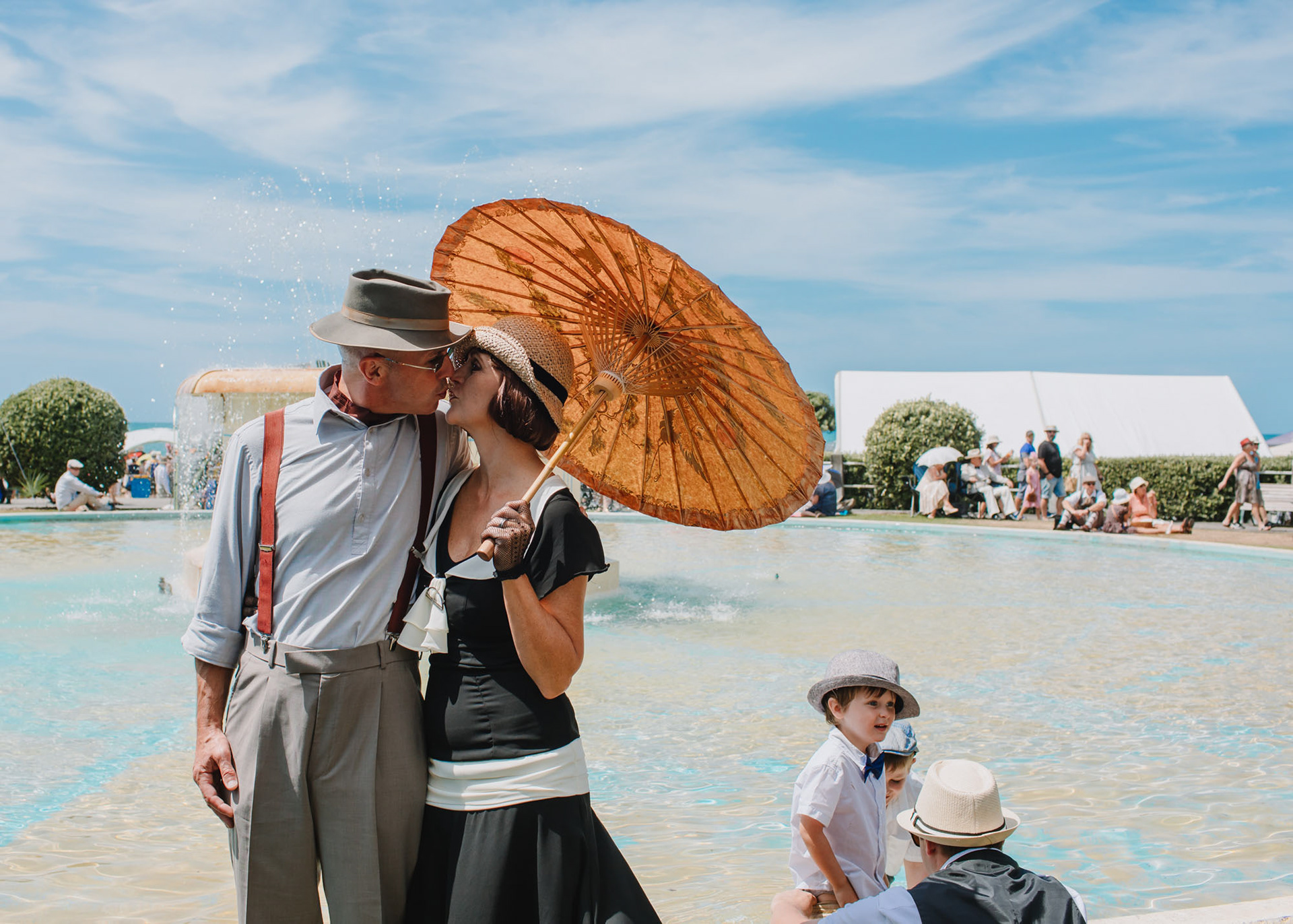 A couple dressed in art deco day wear kiss in front of Tom Parker Fountain