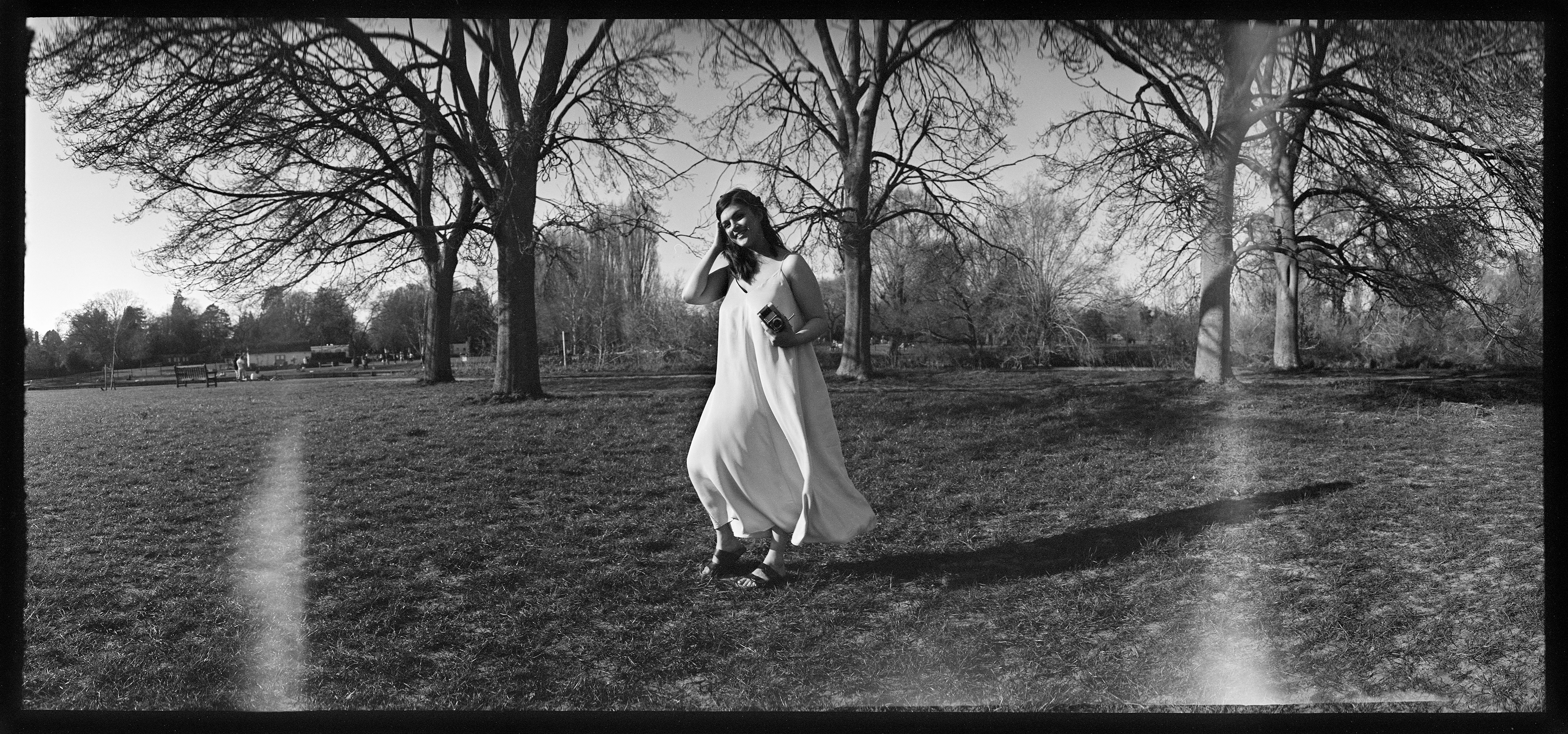 Min is stood in a grassy clearing holding a Voigtländer Vitessa and is wearing a light dress that is being gently buffeted in the wind. She is smiling and brushing her hair behind her ear with her free hand. There are four dark, leafless trees behind her.