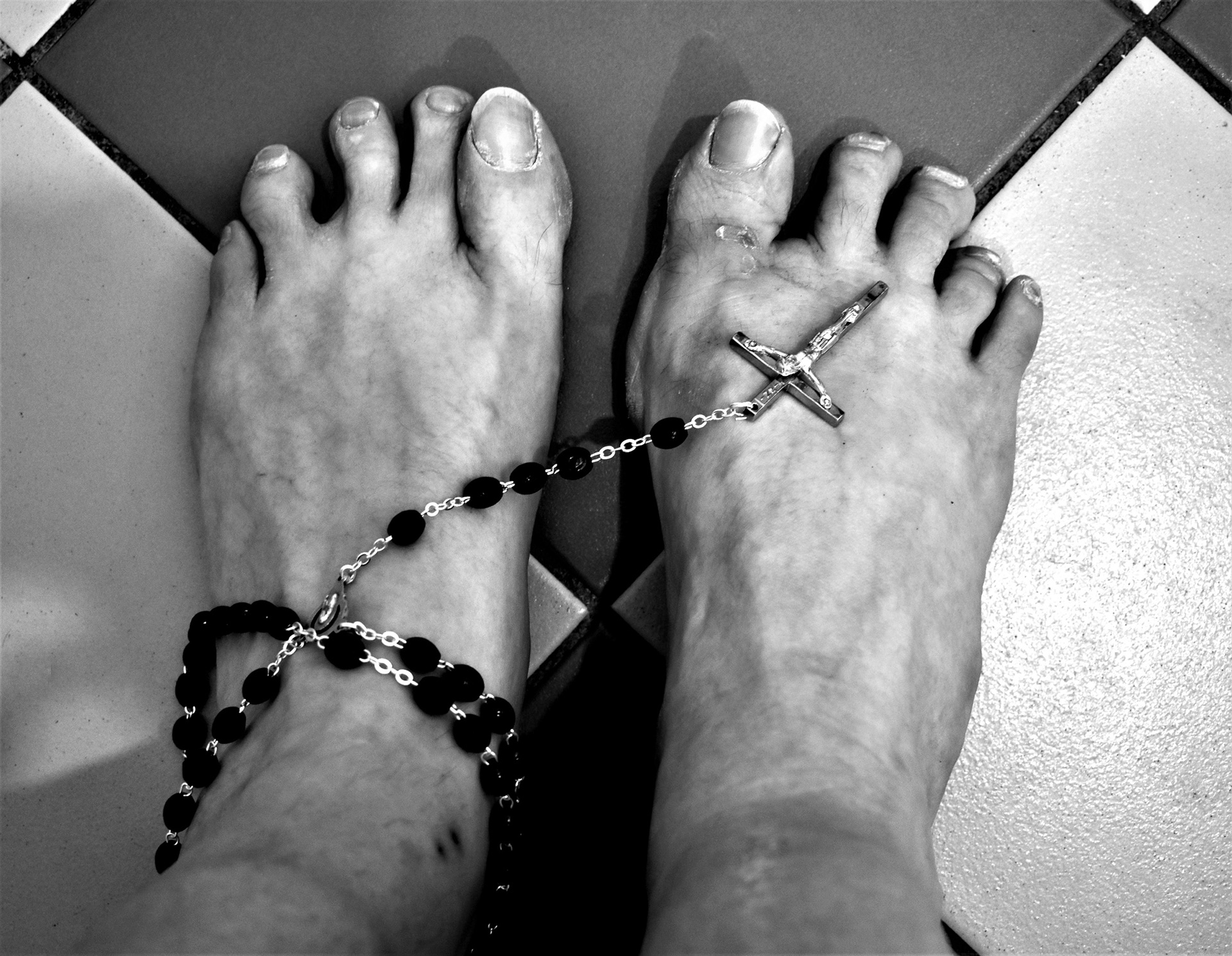 A close up of Kerry's feet, in black and white in high contrast, with mishapen nails and a black rosary draped around them