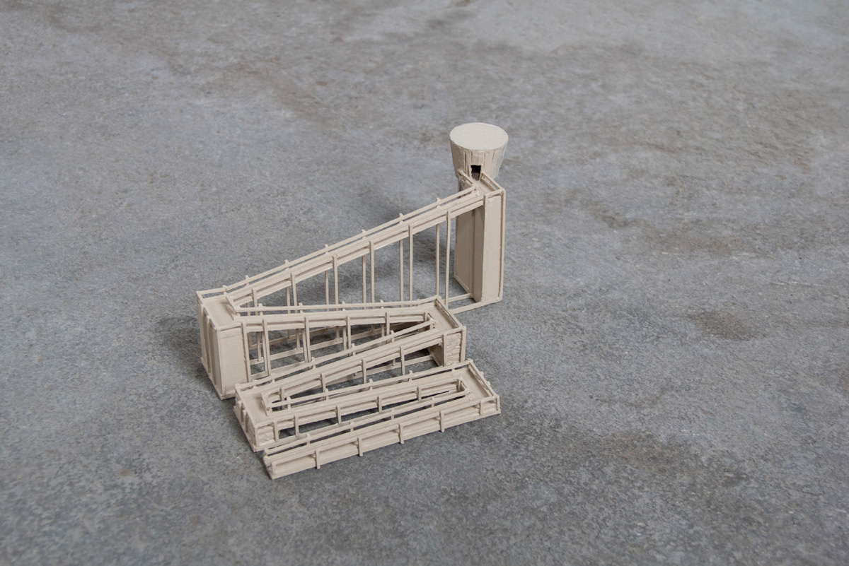 A close-up of a small balsa wood model of a watchtower on top of a concrete floor. It has a long accessibility ramp reaching to the top.