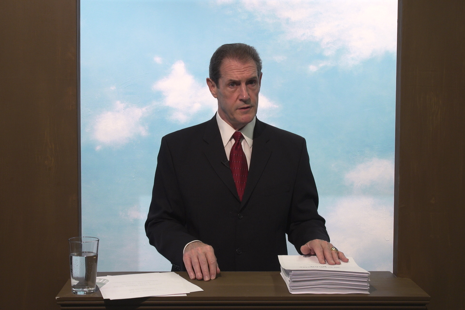 An image still from the short film "Man Above the Fog." The still shows a middle-aged man in a dark blue suit and red tie standing at a podium. Behind him is a wall painted with a blue sky and fluffy clouds. The man's left hand rests on a large stack of papers that he is reading from, to his right is a mostly full glass of water.
