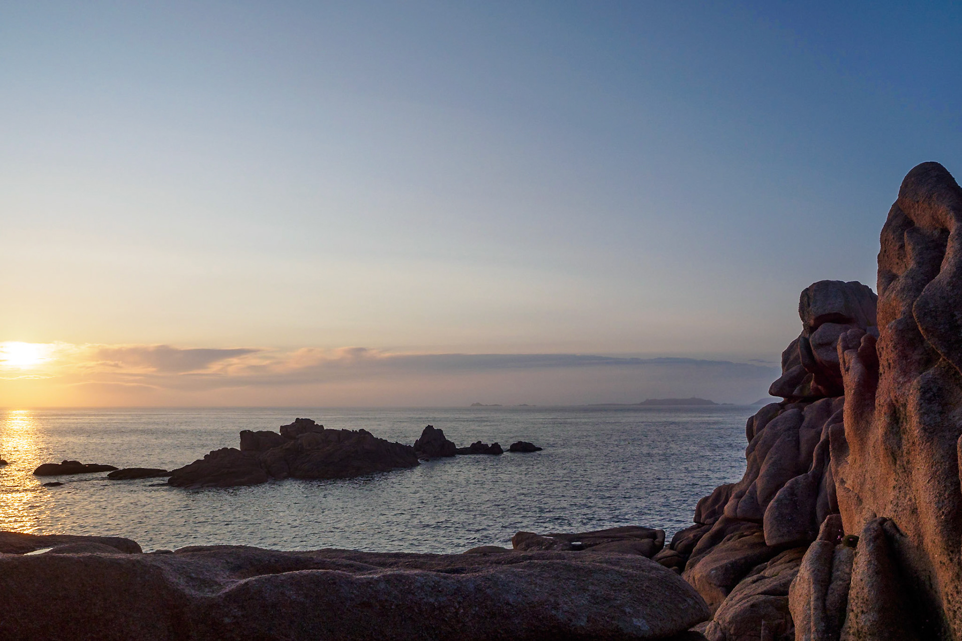The slightly veiled sun setting over the English Channel at Ploumanach, France, reflecting in the water,  casting a soft dim light over the pink granite rocks. A small rock formation in the foreground provides a leading line guiding the eye towards the islands on the horizon.