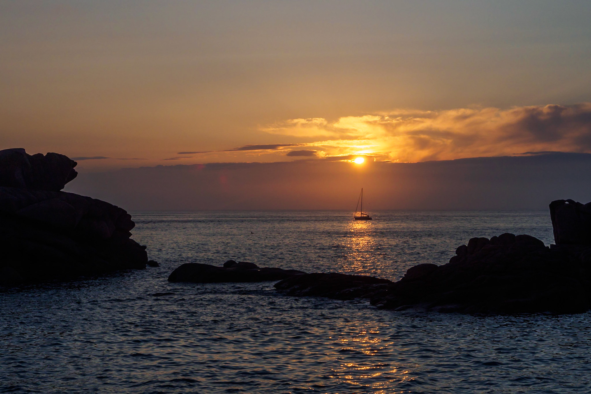 The slightly veiled sun setting over the English Channel at Ploumanach, France, reflecting in the water,  casting a soft dim light over the pink granite rocks. And right in the reflection of the sun on the water, a small boat passing by.